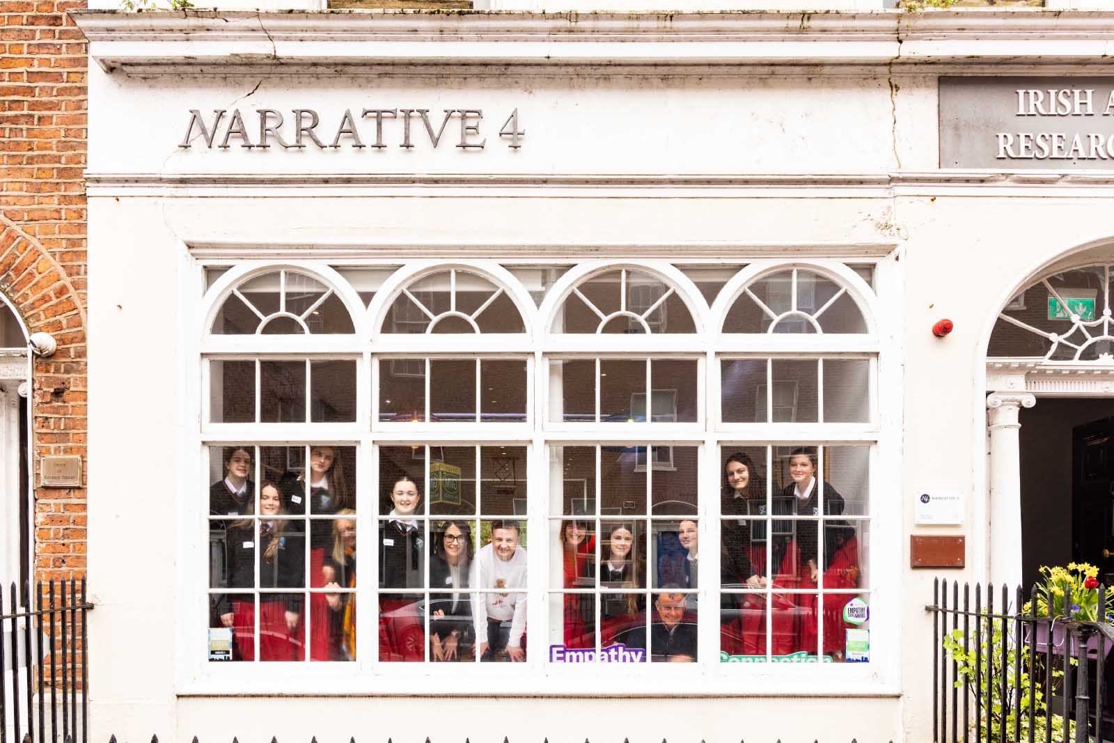 A group of smiling students and adults looking through a window, under the sign Narrative4.