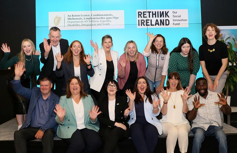 A group of fourteen excited and happy individuals in front of a bright blue background.