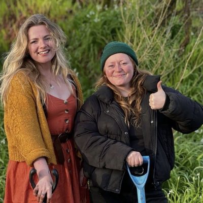 Two people smiling in a field and holding shovels.