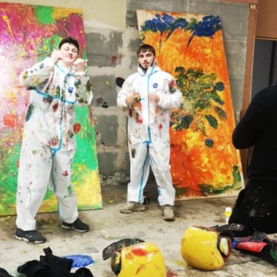 Two people covered in paint splatters, standing in front of painted canvases.