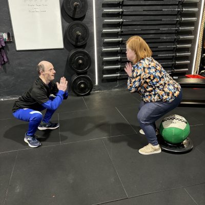 A man and a woman doing squats in a gym. The woman uses a soft ball for support