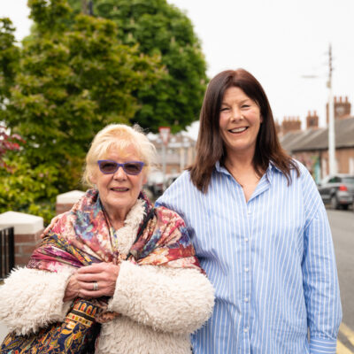 Elderly lady stands beside care worker on residential street as both smile at camera