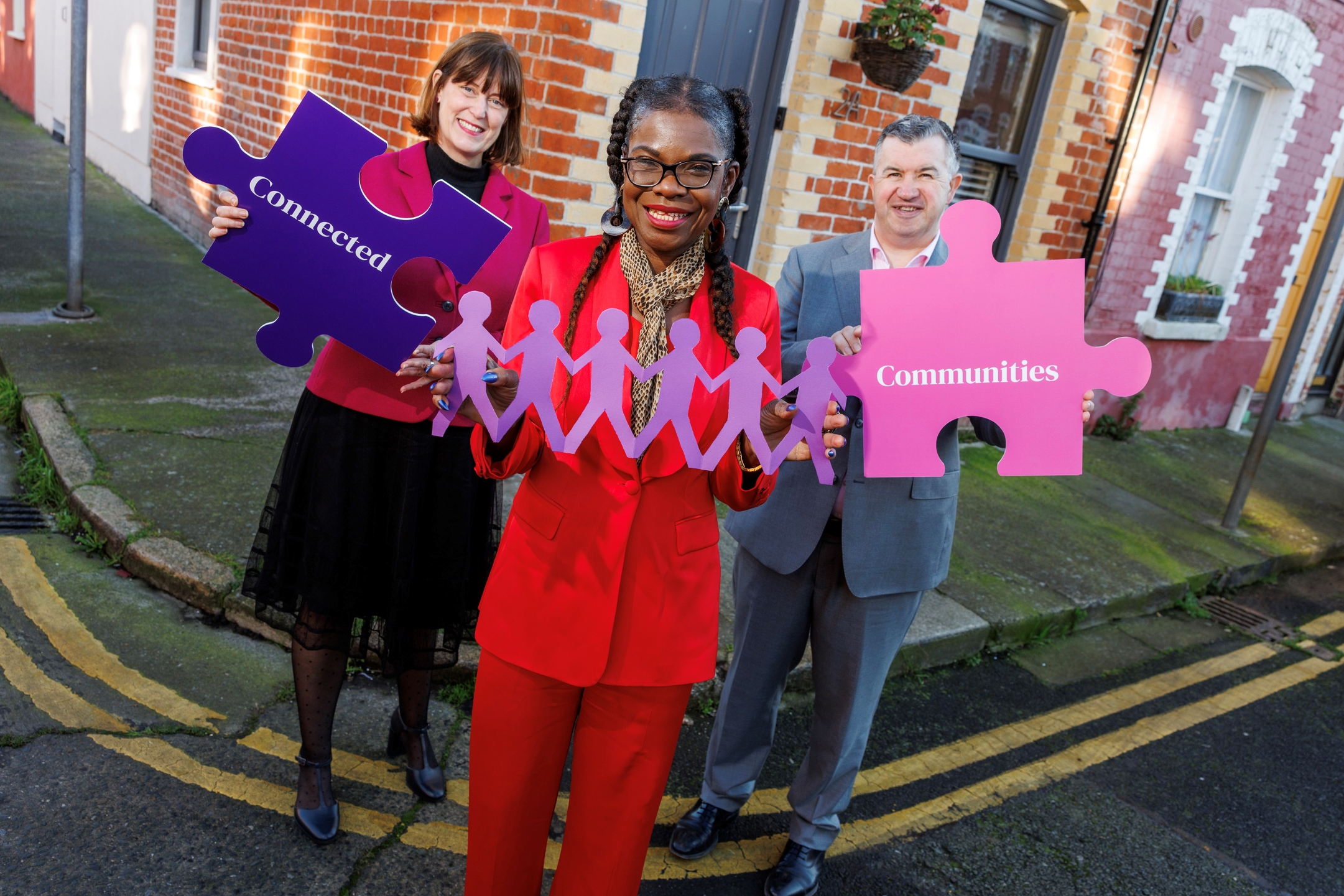 A man and woman hold up jigsaw pieces with the word 'connected' on one and 'communities' on the other, while a third woman holds a paper doll chain of connected people in the middle.