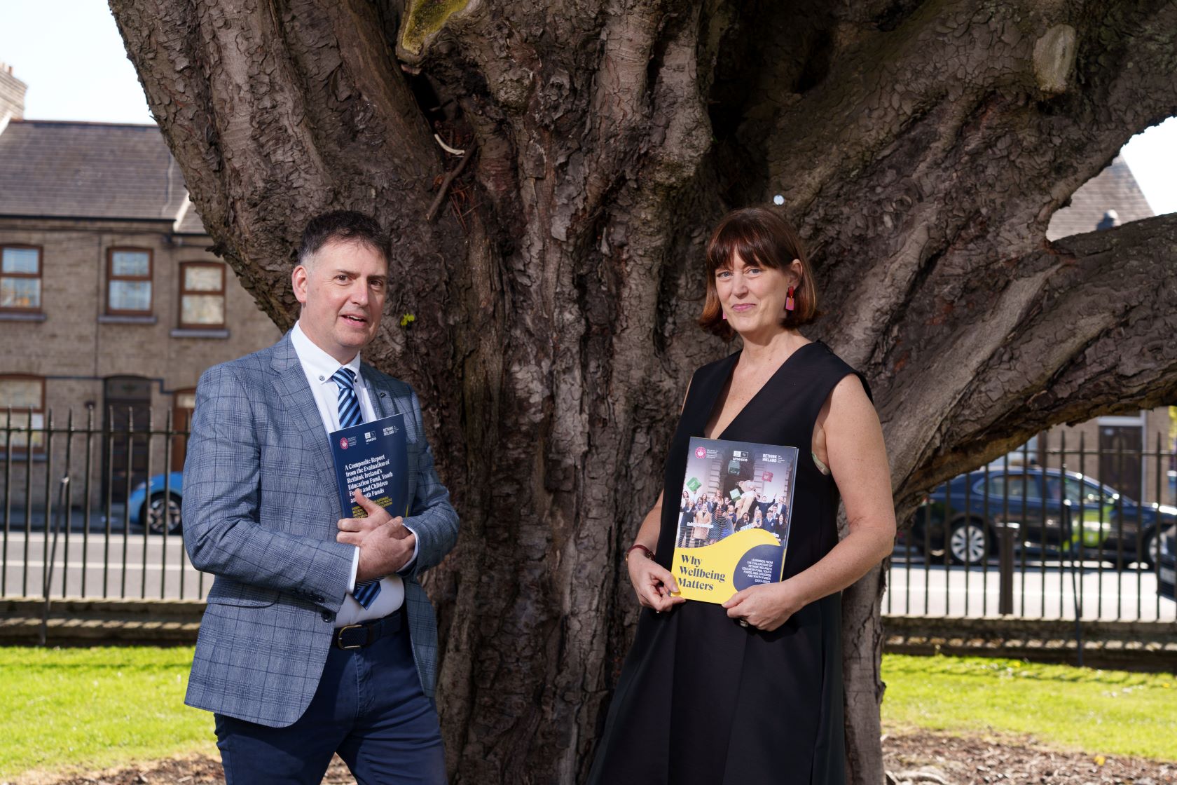 Man and woman hold copy of report in front of tree on a sunny day