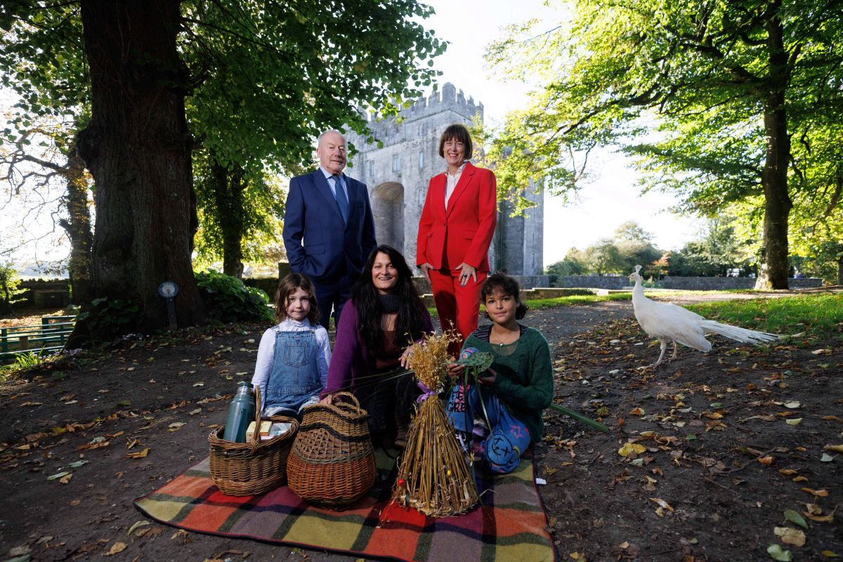 A man and a woman standing with 2 children and a woman kneeling in front on a blanket. A castle is in the background