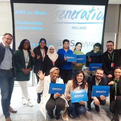 Group of young people and 1 older man holding signs in front of a screen and waving to the camera