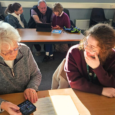 An older woman and a younger girl at a table both looking towards a mobile phone on a table in front of them